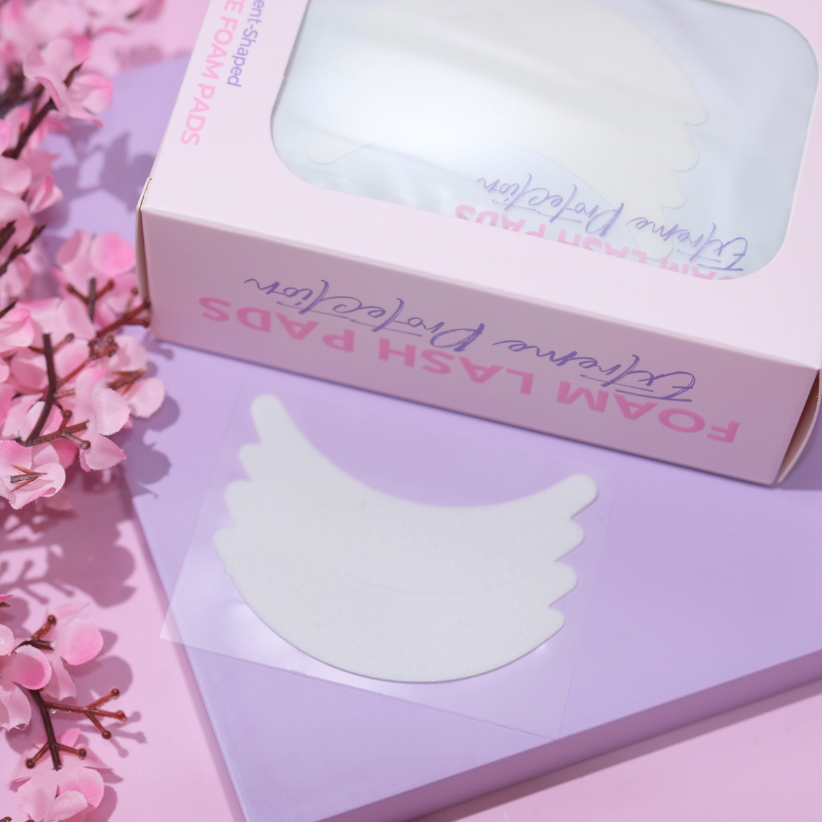 White crescent-shaped foam lash pads displayed on a lavender card next to their pink packaging box, styled with pink cherry blossoms on a pink background.
