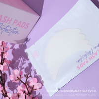 Lash protection pads in a white pouch with a translucent shield graphic, styled on a lavender background with pink packaging and cherry blossom branches. Text reads '50 PAIRS.