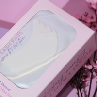 A pink box of foam lash protection pads with a clear window showing white crescent-shaped pads inside, photographed on a pink background with blurred flowers.